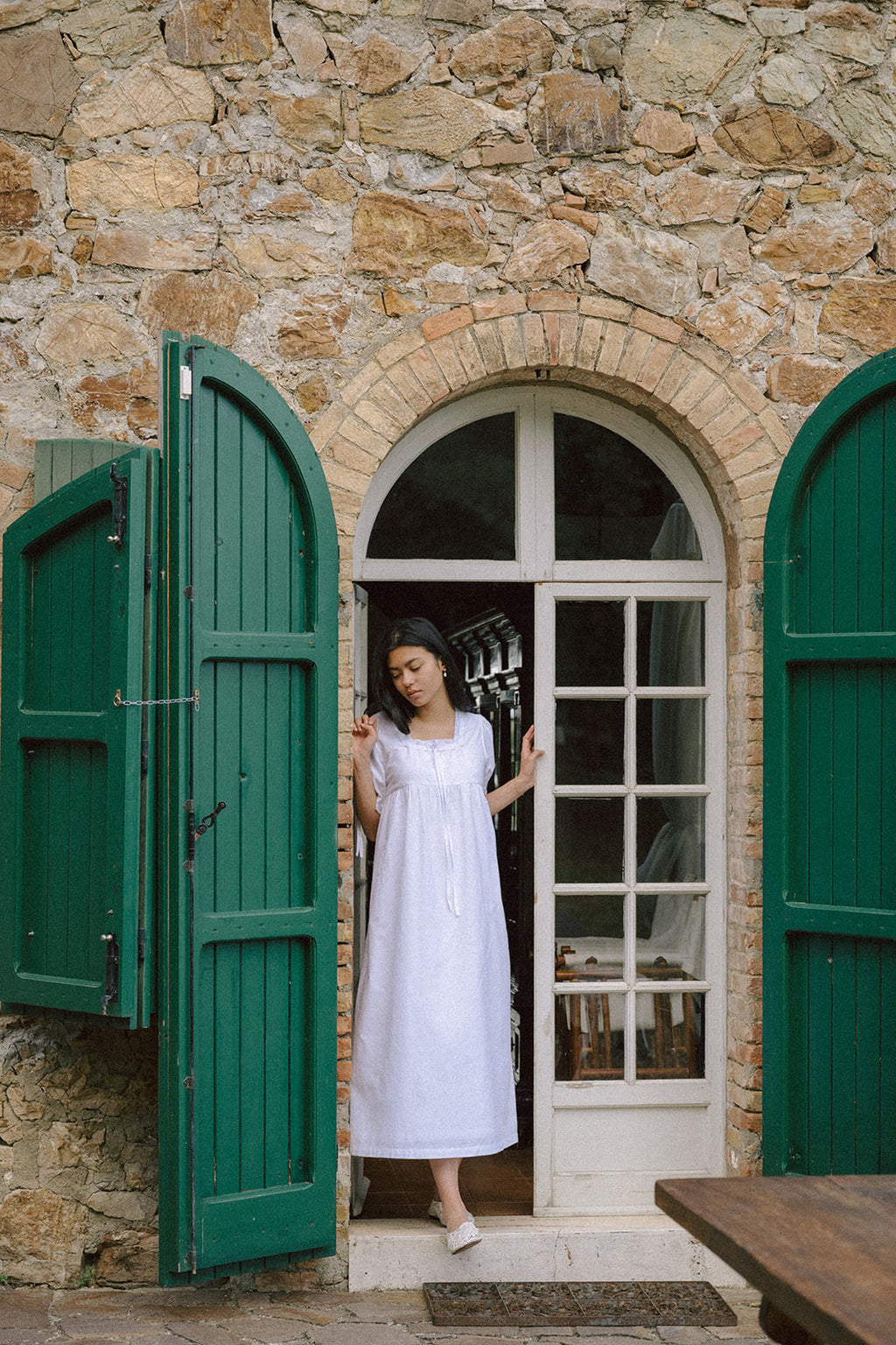 Model wearing the Dace Dress in Salt by Gillian Stevens, standing in a rustic stone doorway with green shutters