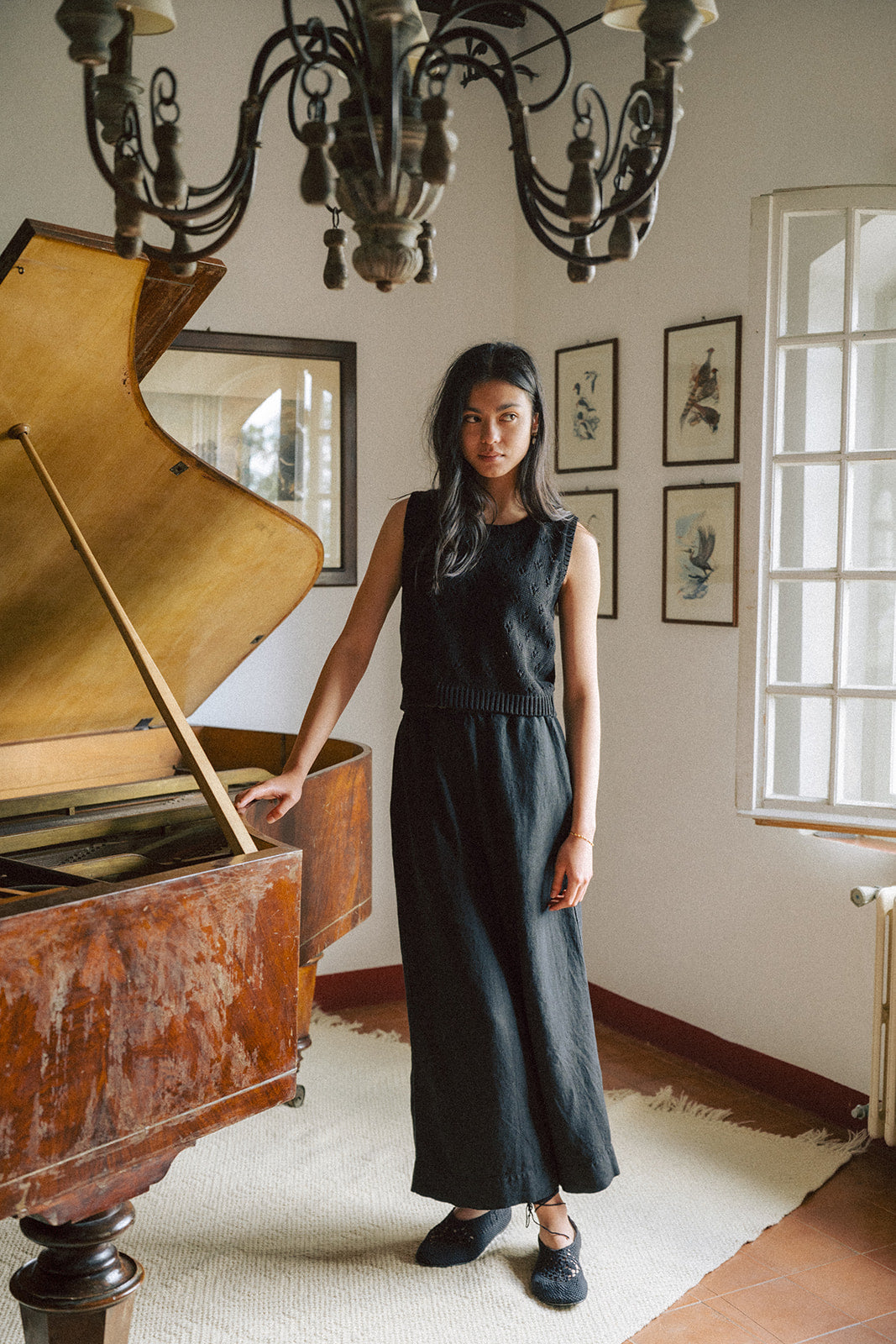 Model wearing the Niah Top in Noir by Gillian Stevens, styled with a black maxi skirt beside a vintage piano