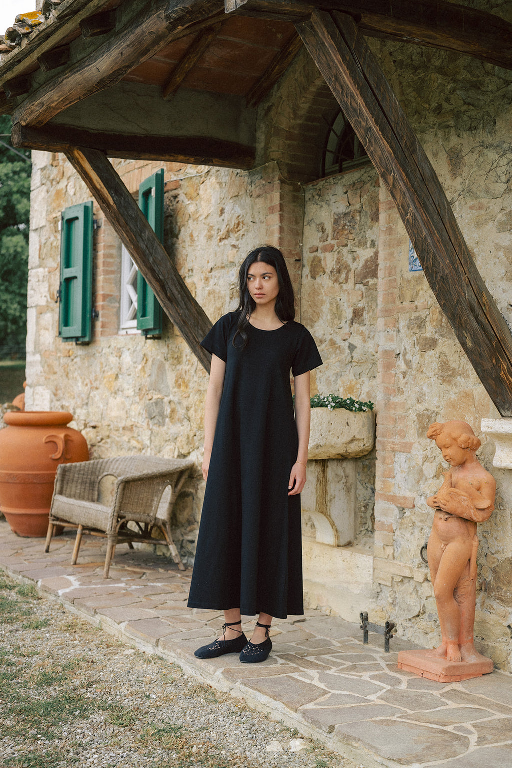 Model wearing the Palmer Dress in Noir by Gillian Stevens, front view beside a rustic stone villa