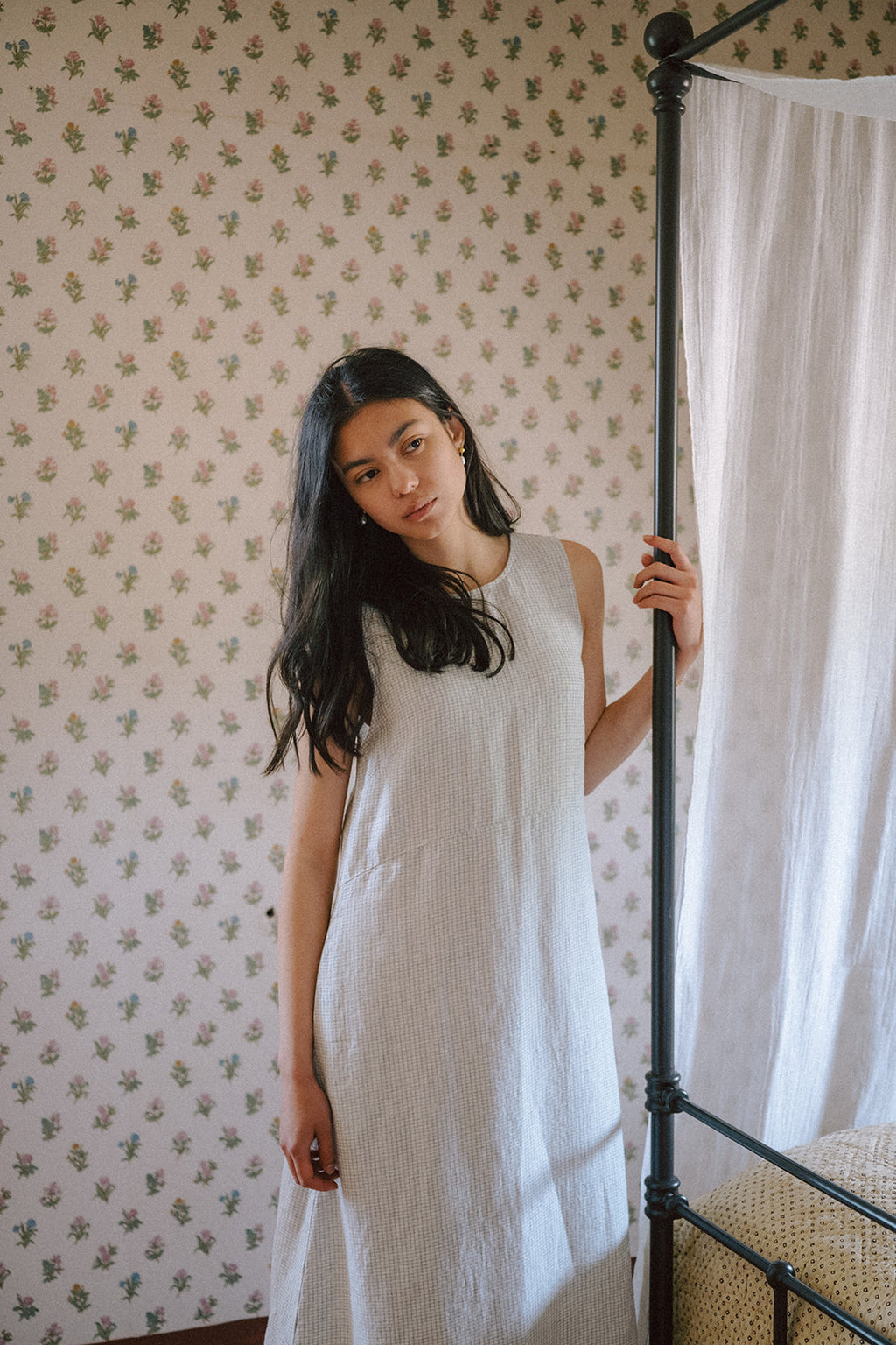 Model wearing the Paige Dress in Dusty Blue Plaid by Gillian Stevens, front view beside an iron bed frame