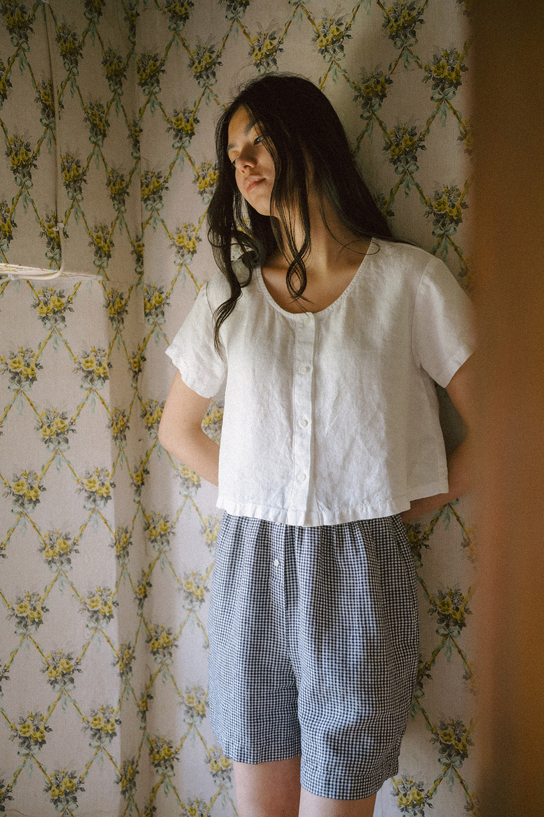Model wearing the Wren Top in Salt by Gillian Stevens, paired with gingham shorts against floral wallpaper