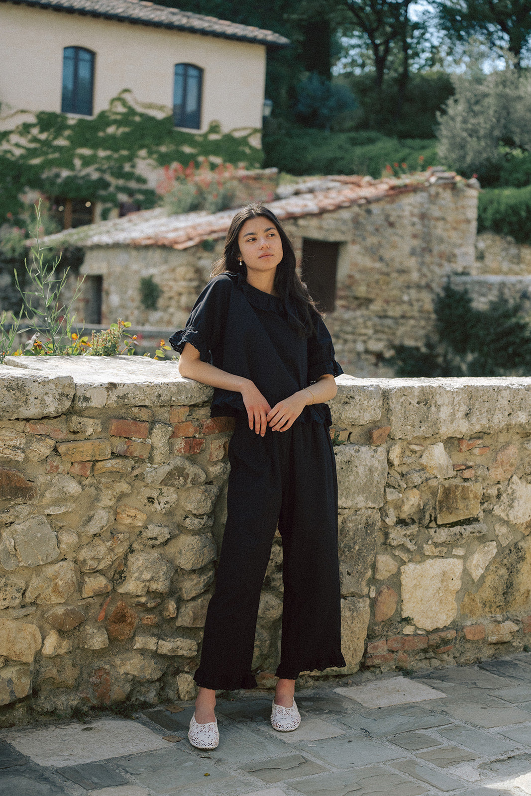 Model wearing the Arlette Pant in Noir by Gillian Stevens, full-length front view against a rustic stone wall