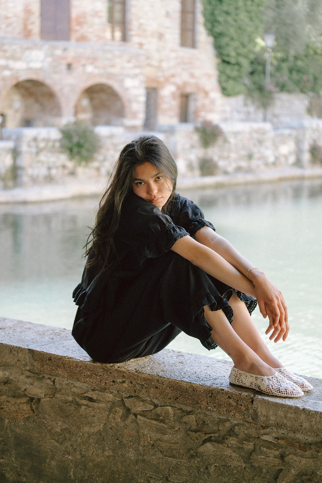 Model seated on stone wall in the Arlette Pant in Noir by Gillian Stevens, styled with a black top, Italian setting