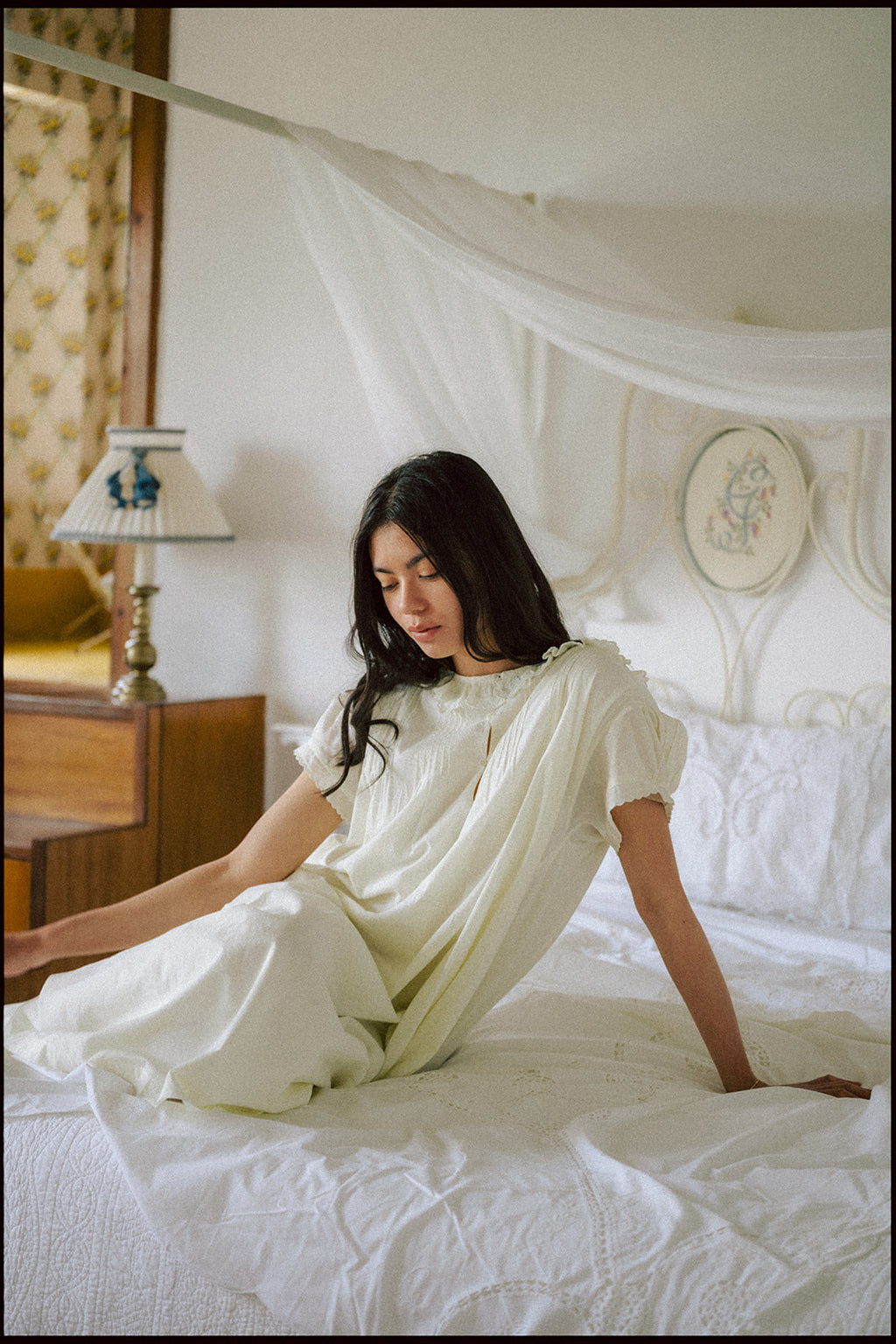 Model seated on a canopy bed wearing the Clover Dress in Butter by Gillian Stevens