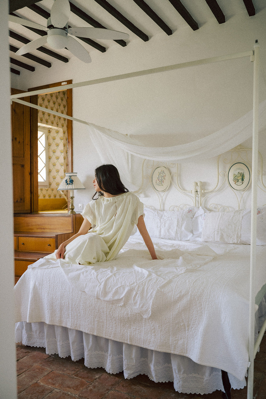 Model seated on a canopy bed in the Clover Dress in Butter by Gillian Stevens, back view in a rustic bedroom setting