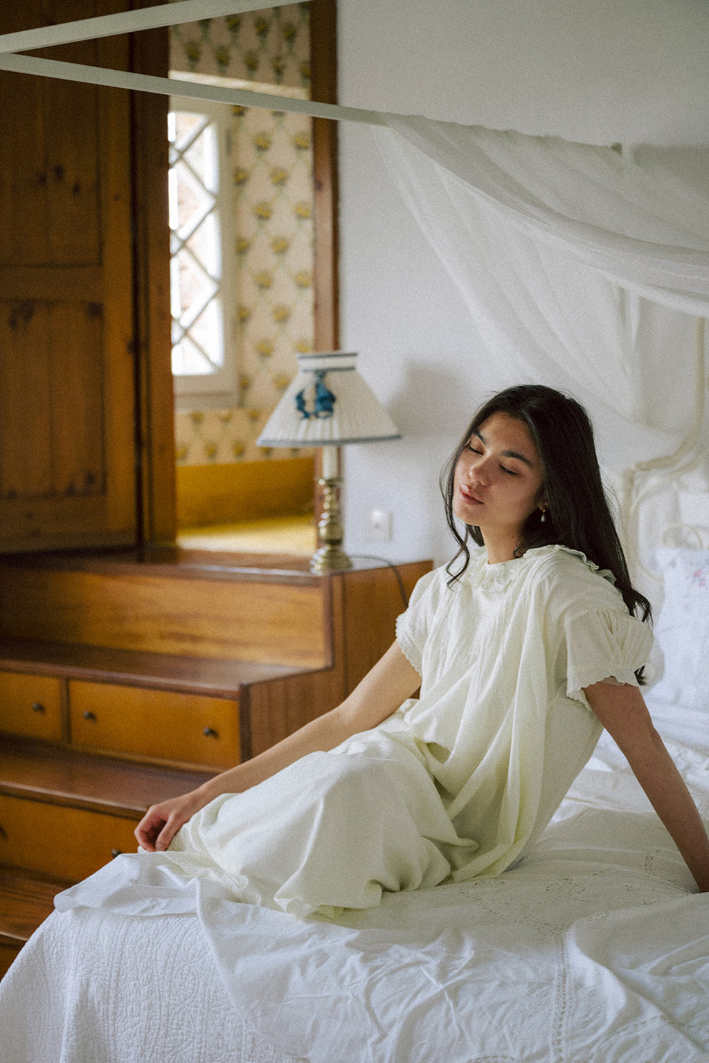 Model seated on a canopy bed wearing the Clover Dress in Butter by Gillian Stevens, front view