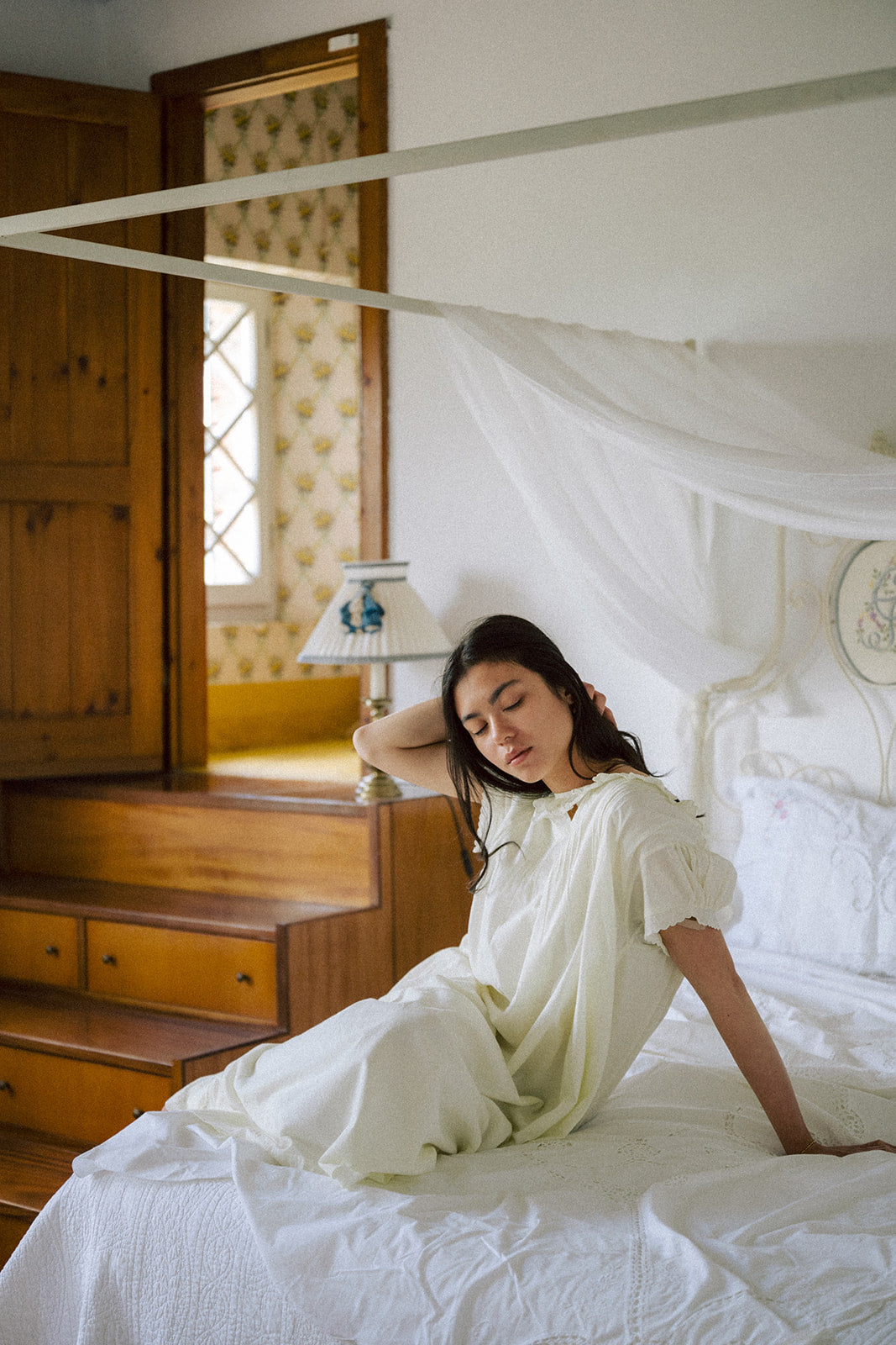 Model seated on a canopy bed wearing the Clover Dress in Butter by Gillian Stevens
