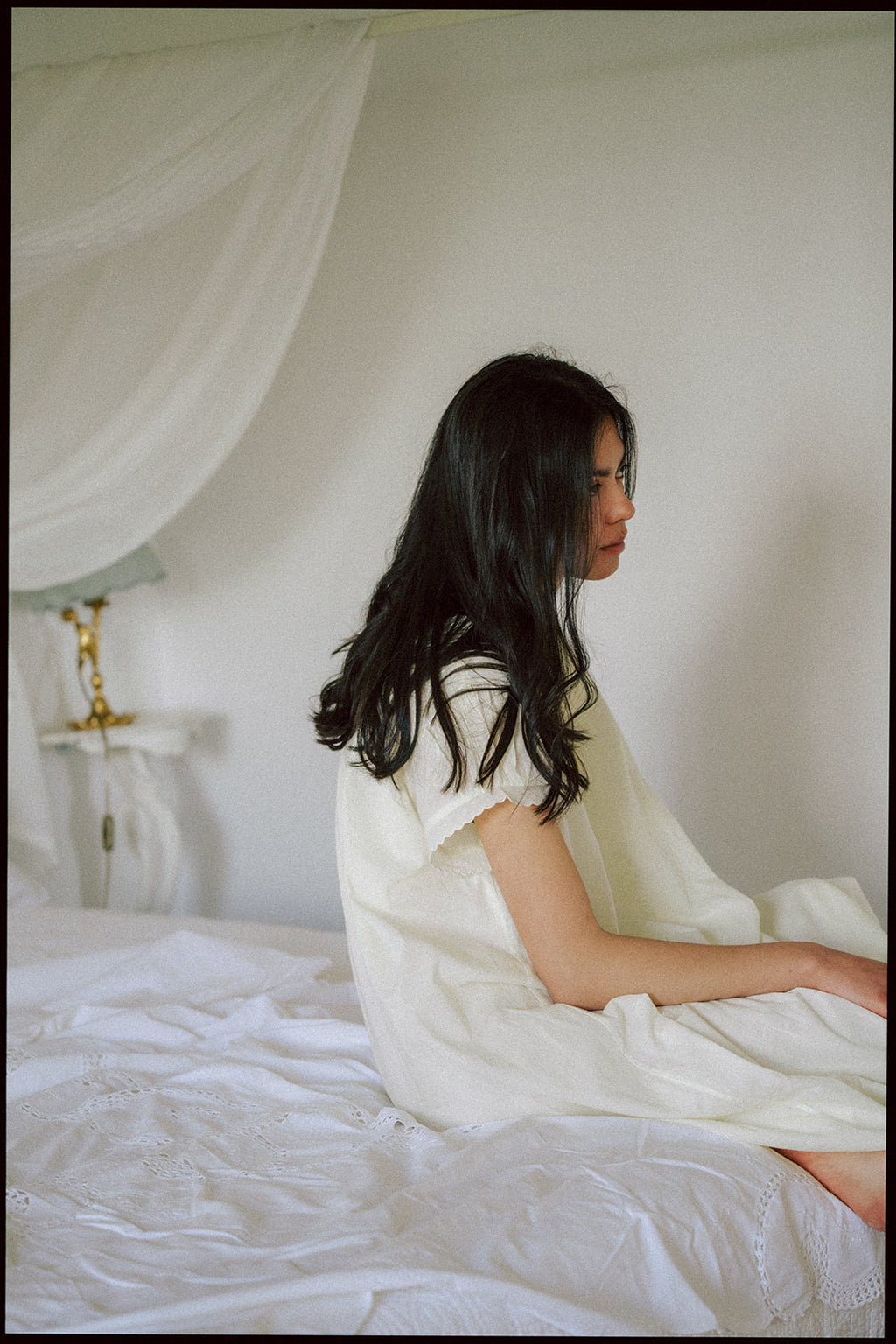 Model seated on a bed wearing the Clover Dress in Butter by Gillian Stevens, side profile view