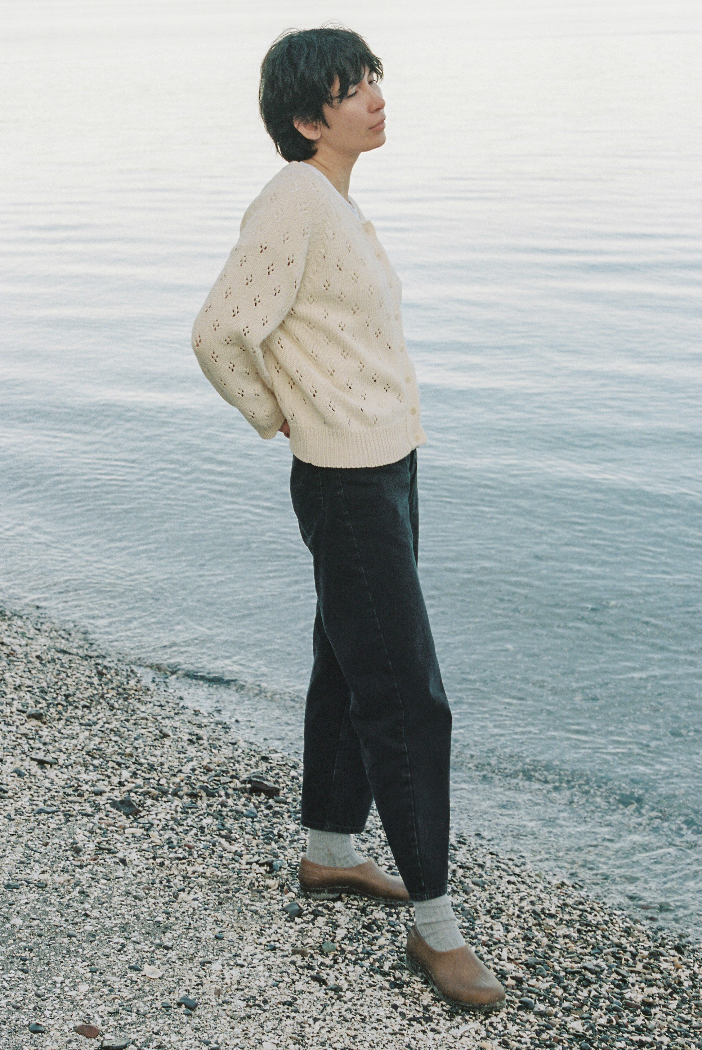 Model wearing the Dara Jean in Noir by Gillian Stevens, side view at a pebbly waterfront
